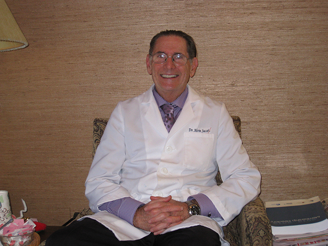 The image shows a man sitting in a chair, wearing a white lab coat, with glasses and a beard, smiling at the camera.