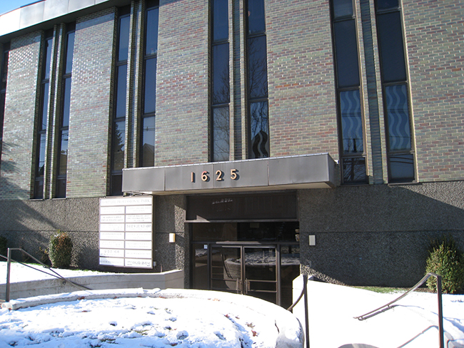 The image shows a building with a brick facade, a large glass door entrance, and a sign above the door displaying an address.