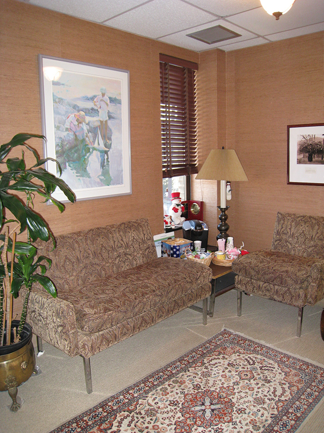 The image shows a well-lit office space with a comfortable sitting area featuring a couch, chair, coffee table, potted plant, and framed artwork on the wall.