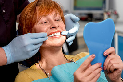 A woman sitting with her mouth open while holding a blue dental impression tray, being attended to by a dental professional during a teeth cleaning appointment.