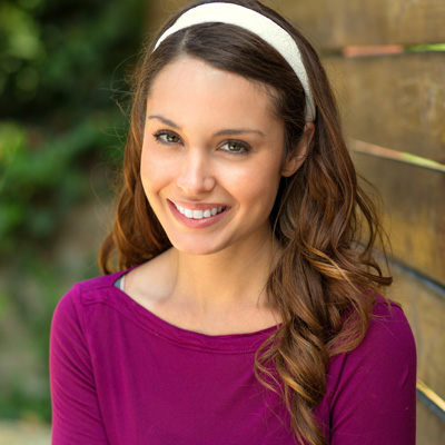 A smiling woman with long hair and a headband, wearing a purple top, stands against a wooden wall.