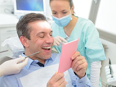 The image shows a man sitting in a dental chair with a smile on his face, holding up a pink card or booklet, surrounded by dental professionals who appear to be engaged in a discussion or examination.