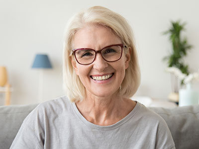 The image shows a woman with short blonde hair, wearing glasses and a light-colored top, smiling at the camera. She has a pleasant expression and appears to be indoors with a homey background that includes a lamp and decorative items.