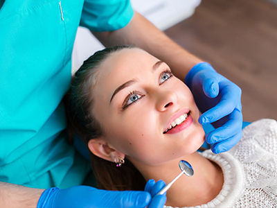 The image shows a young woman with blue eyes receiving dental care from a professional wearing a surgical mask and cap, who is adjusting her teeth with a tool while she smiles.