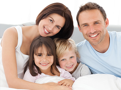 A family of four, including two adults and two children, smiling together on a bed with a white sheet.