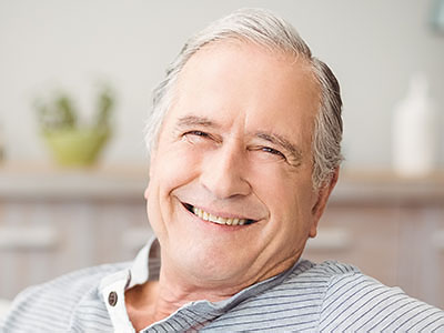 The image shows an older man with gray hair, smiling at the camera while sitting comfortably indoors.