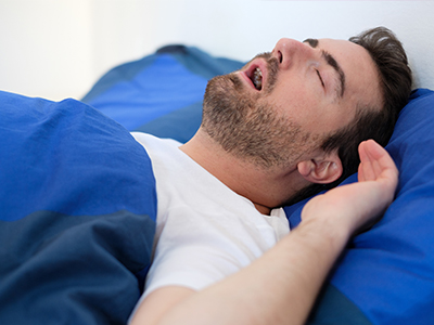 The image shows a man lying in bed with his eyes closed, appearing to be asleep or resting, with a blue comforter on the bed.