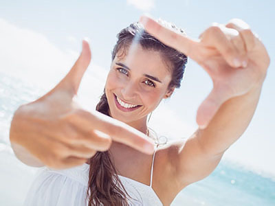 A young woman with long hair smiles while holding up her index finger to frame a picture of herself against a clear sky background.